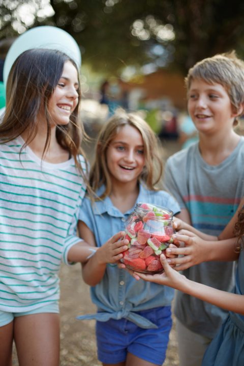 Three children stand outdoors at Wildflower Ranch near Fort Worth TX, smiling and holding a glass container filled with assorted candies and gummies.