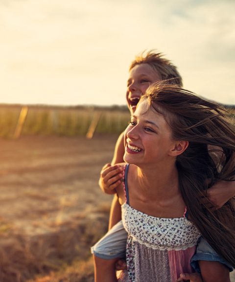 Two children outdoors at sunset in Wildflower Ranch, one giving the other a piggyback ride. Both smile with wind in their hair and a field behind them, capturing the joy of life near new construction homes Fort Worth families love.