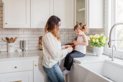A woman stands in a bright kitchen next to a young girl sitting on the counter, holding a peeled orange. Sunlight fills the space of their Dallas Fort Worth home, and a vase of yellow tulips sits by the window.