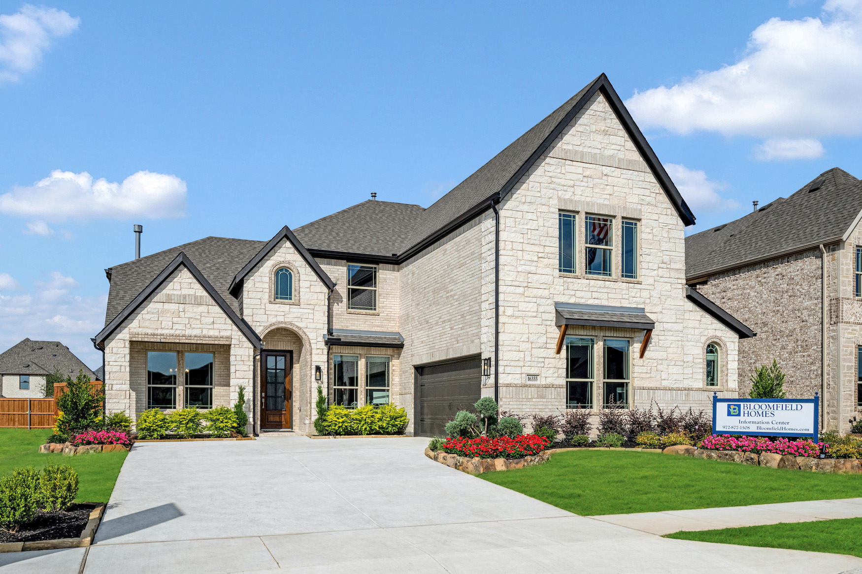 Two-story stone house with a driveway, manicured lawn, landscaped flowerbeds, and a "Blackwell Homes" sign in the front yard under a blue sky with scattered clouds.