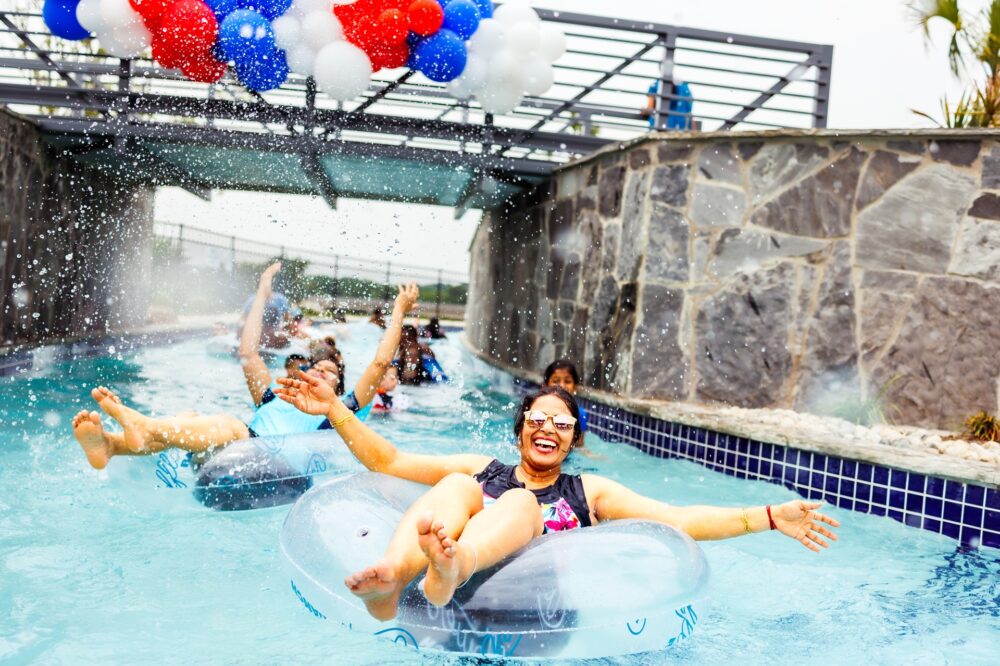 People riding on inflatable tubes in a lazy river, smiling and splashing under a bridge decorated with red, white, and blue balloons at Wildflower Ranch—one of the most exciting new homes for sale communities near Fort Worth TX.