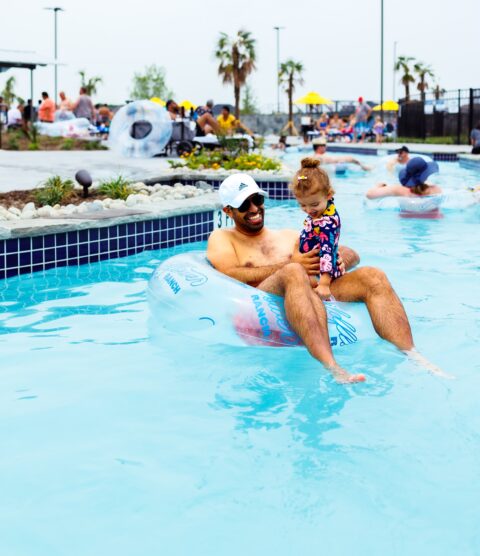 A man wearing sunglasses and a white cap sits on a pool float holding a young child in a colorful swimsuit at the Wildflower Ranch community pool, surrounded by other families enjoying the amenities of these new construction homes in Fort Worth.